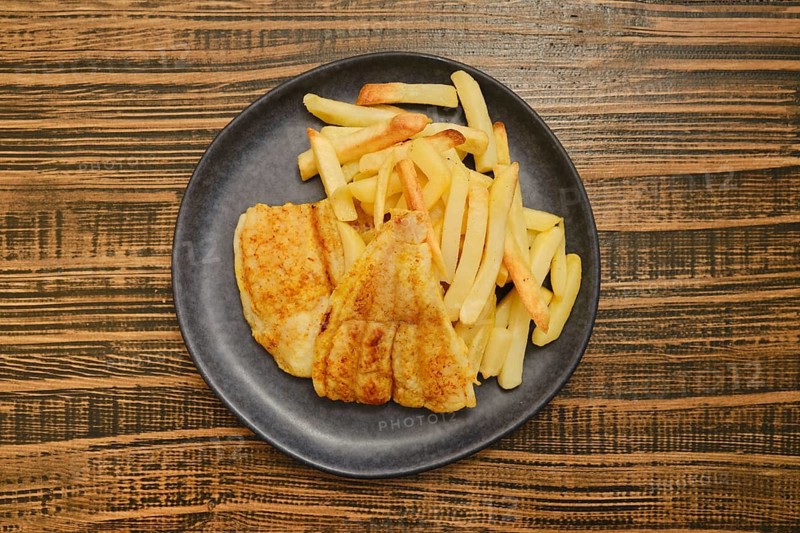 A plate features a crispy fried fish fillet alongside a generous portion of golden french fries. The meal is set on a textured wooden table, inviting a casual dining experience