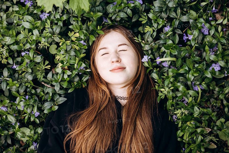 A thoughtful teenage girl lies on a lush bed of green periwinkle leaves and violet blossoms, surrounded by nature in a peaceful and reflective moment outdoors