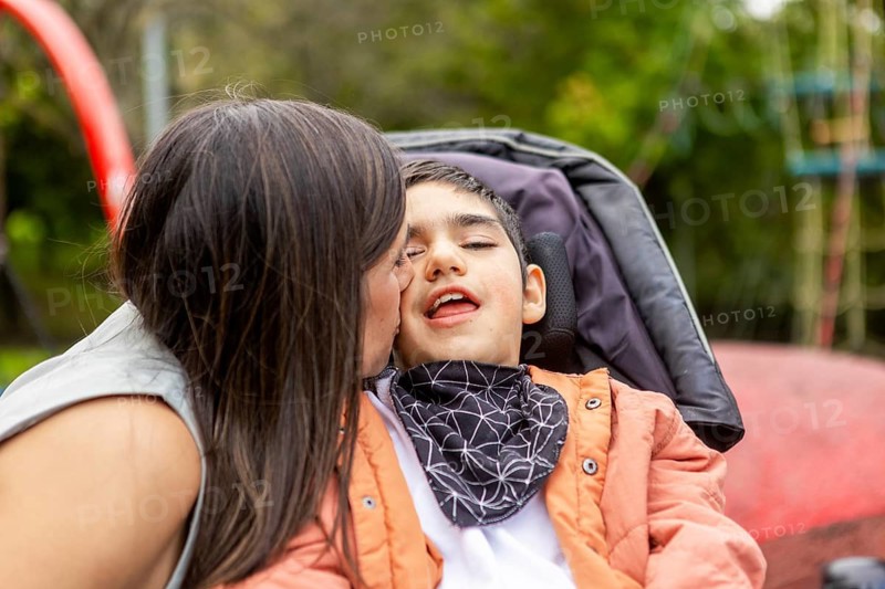 A tender moment as a mother kisses her son with cerebral palsy on the cheek. The son, seated in a wheelchair. Greenery in the background evokes a serene playground setting