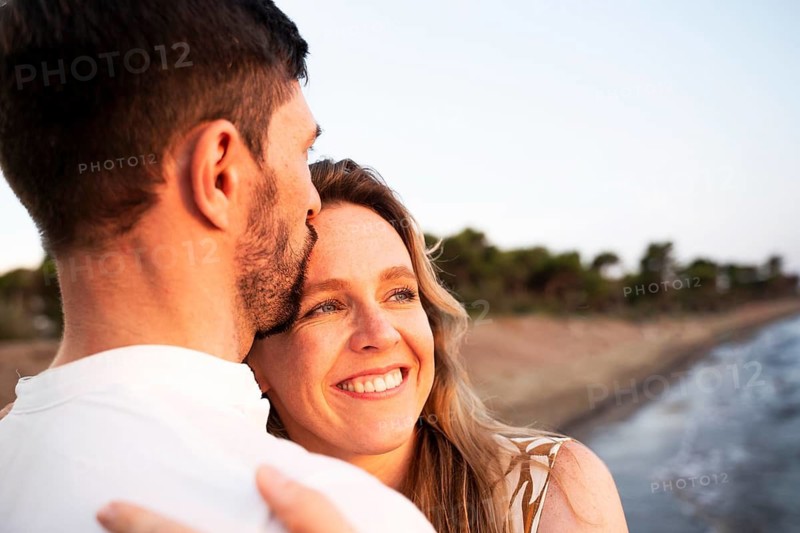 A loving couple embraces on an Italian beach at sunset, their smiles reflecting happiness and togetherness. The serene coastal backdrop enhances the romantic atmosphere