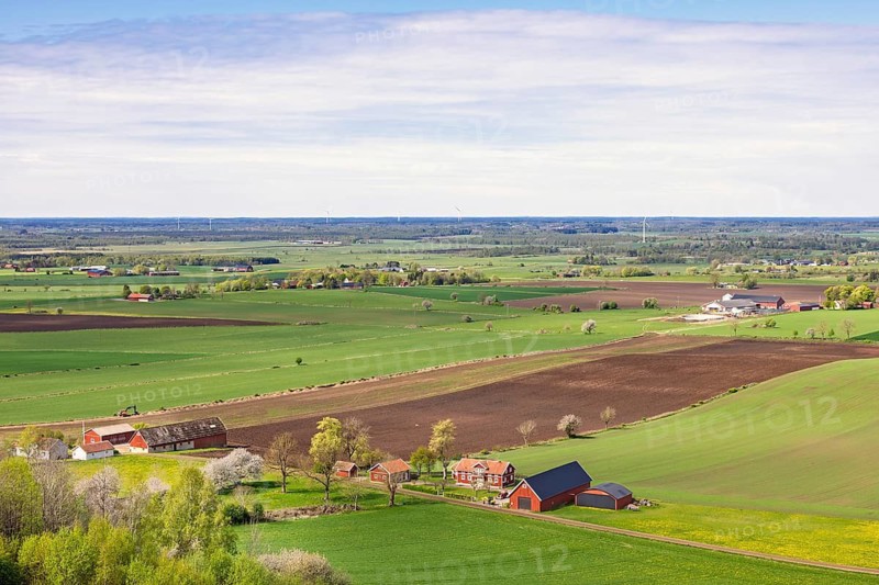 Wide rural landscape with extensive green and brown agricultural fields stretching toward the horizon. Scattered farmhouses and barns in red and white sit among trees and grassy areas in springtime, Sweden
