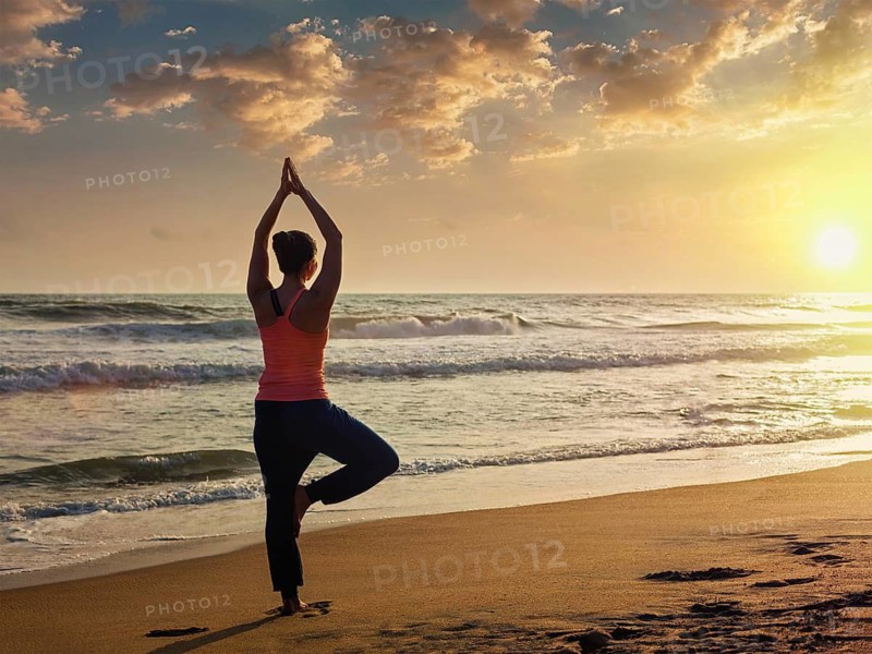 Yoga outdoors, sporty fit woman doing Hatha yoga asana Vrikshasana tre pose on tropical beach on sunset