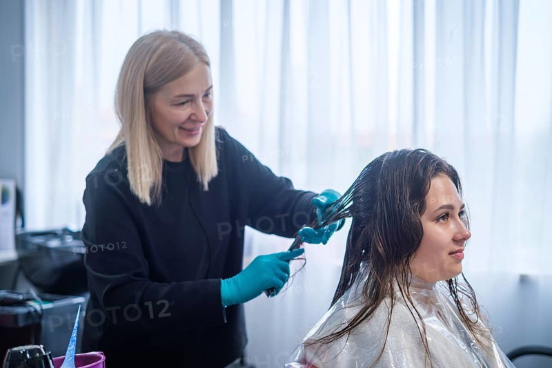 A hair stylist applies keratin treatment to a client's hair in a beauty salon. The stylist wears gloves and carefully works on the hair. The setting has large windows allowing light in