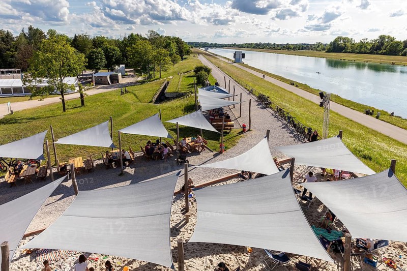People relaxing under awnings by a river running through a green park, Oberschleißheim regatta course