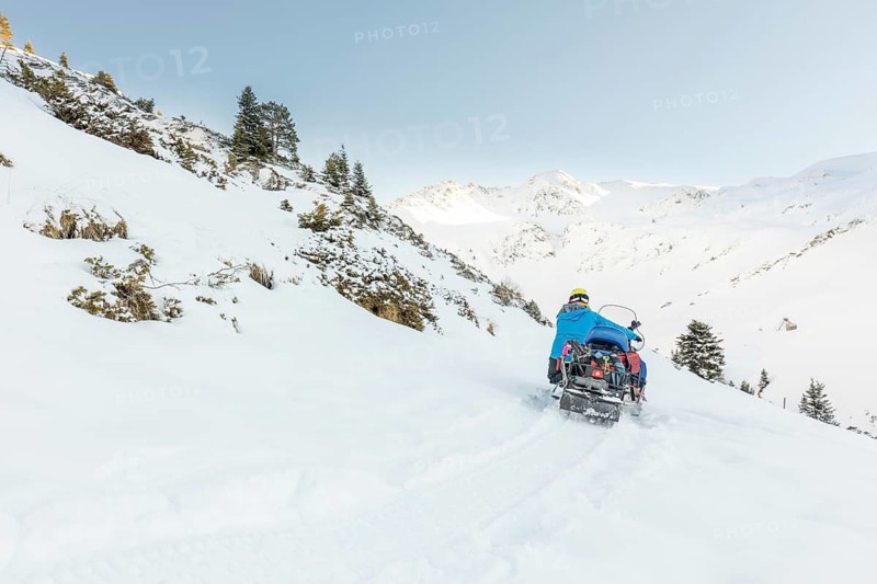 A man in winter gear drives a snowmobile through a pristine, snow-covered mountainous landscape. The blue sky and distant peaks create a breathtaking backdrop for adventure