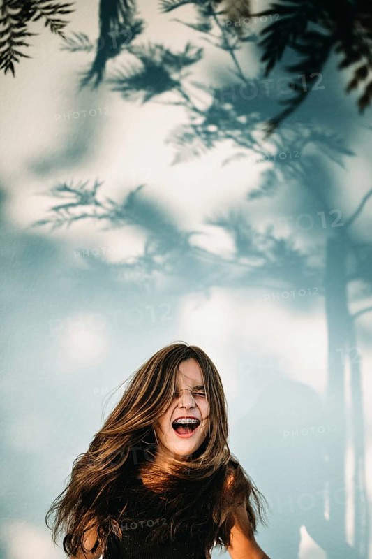 A teen girl with long hair and braces is joyfully smiling. Soft shadows of tree leaves create an artistic backdrop, enhancing a light, playful mood