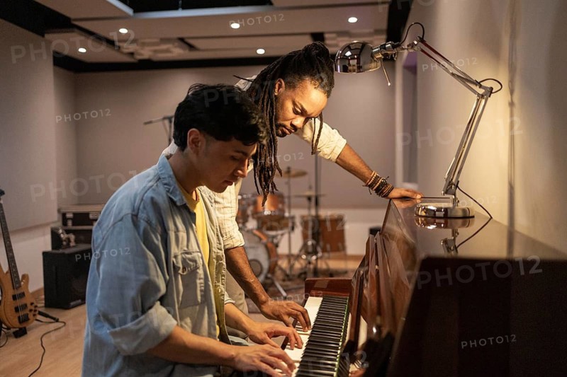 Two musicians work together at a piano, collaborating creatively in a modern recording studio. Instruments and equipment are in the background, highlighting the music-making process