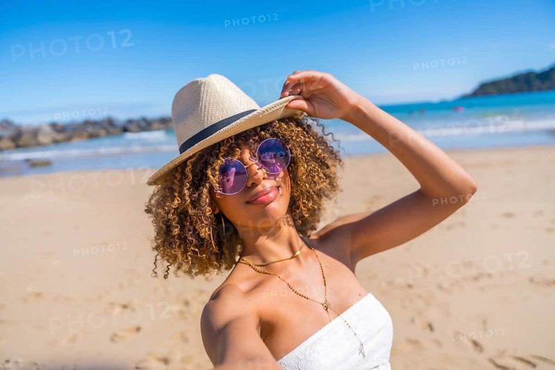 Personal perspective of a beauty young latin woman holding a sun hat attached at her head while taking selfie a beach