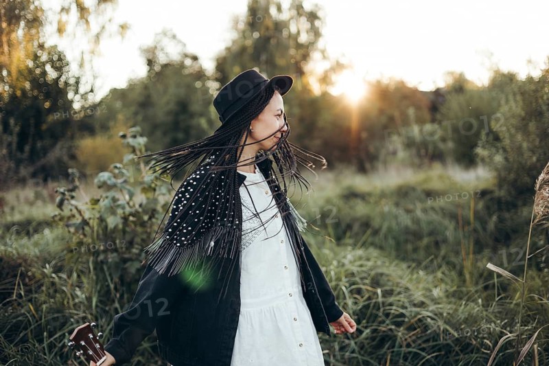 A woman stands in a serene field at sunset, wearing a hat and white dress. Her long braids dance in the breeze as the golden sun sets behind distant trees, creating a peaceful ambiance