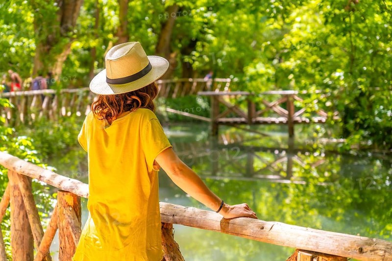 A woman in a yellow shirt is standing on a wooden bridge looking at the water. The scene is peaceful and serene, with the woman enjoying the view and the natural surroundings
