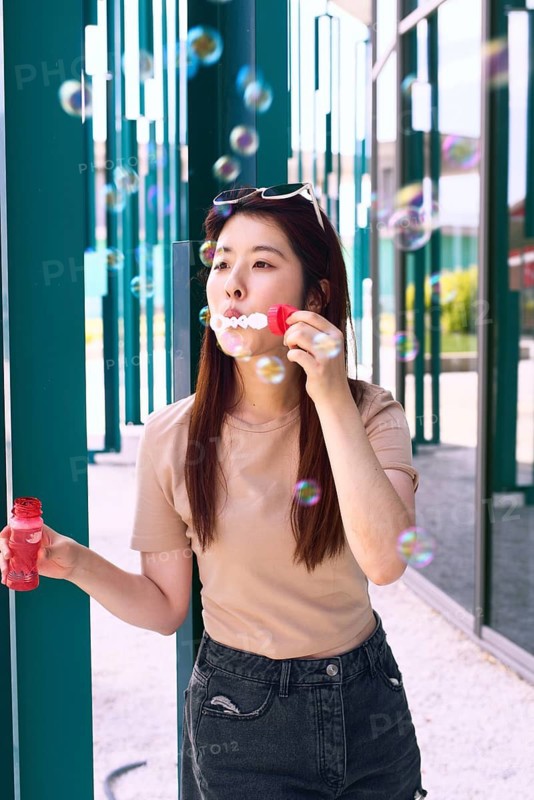 A young woman enjoys a sunny day by blowing bubbles outdoors. She is wearing sunglasses and a casual outfit, surrounded by the playful reflections of bubbles floating around