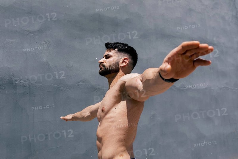 Muscular man with outstretched arms contrasts against a plain gray wall, embodying strength and balance. His posture and physique emphasize dedication to fitness and sport