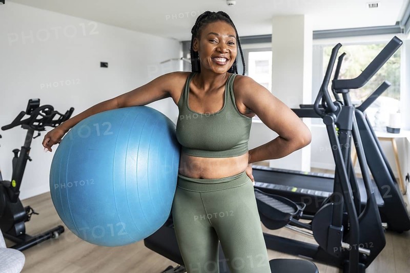 Happy woman wearing sportswear, leaning on a fitness ball in a hotel gym, promoting a healthy lifestyle during business trips