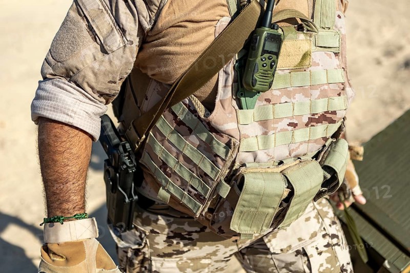 Close up of a man wearing a tactical vest in a desert environment. The camouflage gear is equipped with a radio, highlighting readiness and communication efficiency