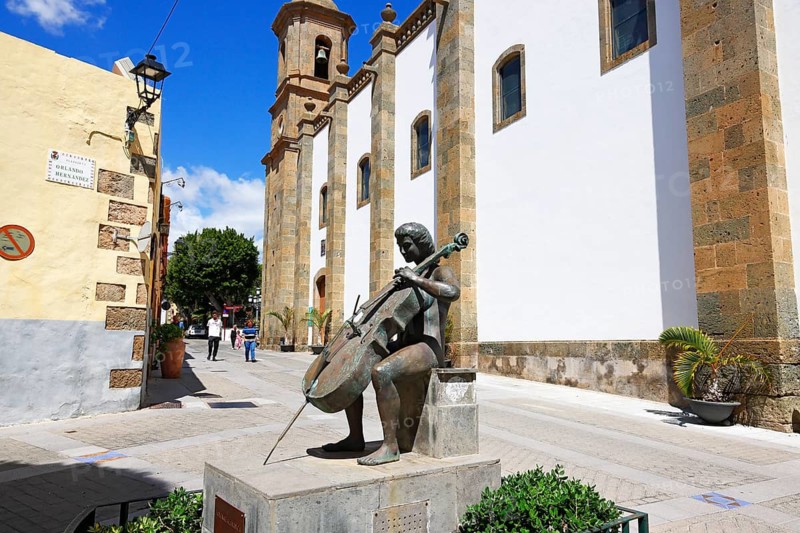 Bronze figure of a cellist Homenaje a la Musica by A. L. Benitez next to the cathedral Iglesia de San Sebastian in the historic centre of Agüimes, Gran Canaria, Canary Islands, Spain