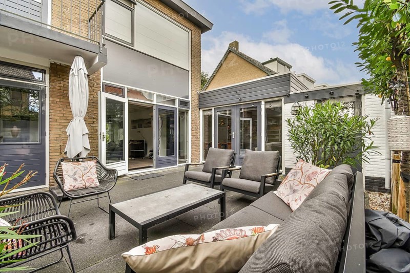 A modern patio featuring comfortable outdoor seating with cushions and a central table, surrounded by greenery and architectural elements. Ideal for relaxation or gatherings