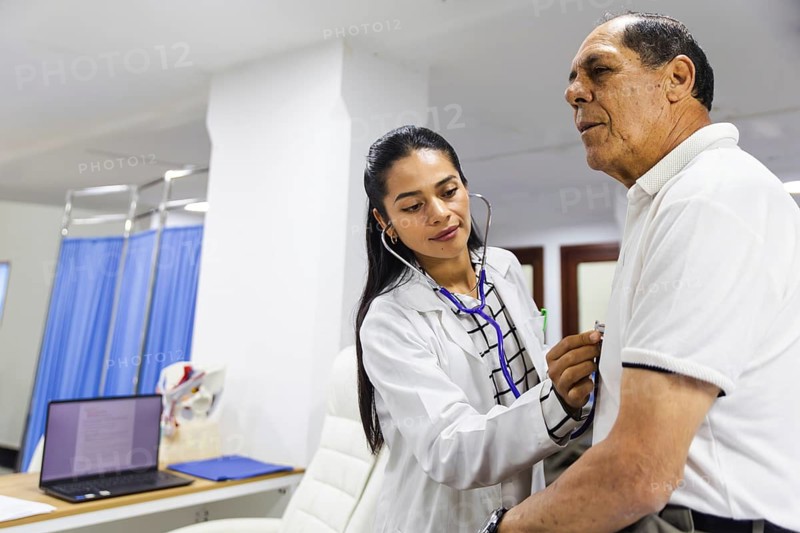 A young female doctor attentively examines an elderly male patient with a stethoscope in a Latin American hospital, showcasing professional healthcare in a clinical setting
