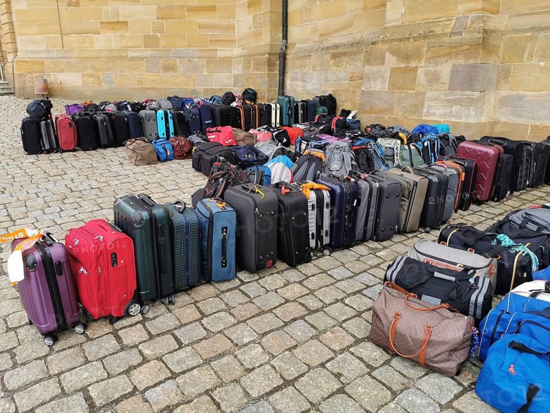 The luggage of a group of travellers stands in rows on the pavement, outside, suitcases and bags