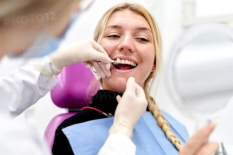 A dentist wearing gloves examines a smiling patient in a modern dental clinic. The patient holds a mirror while the dentist checks her teeth, highlighting the clean, bright setting