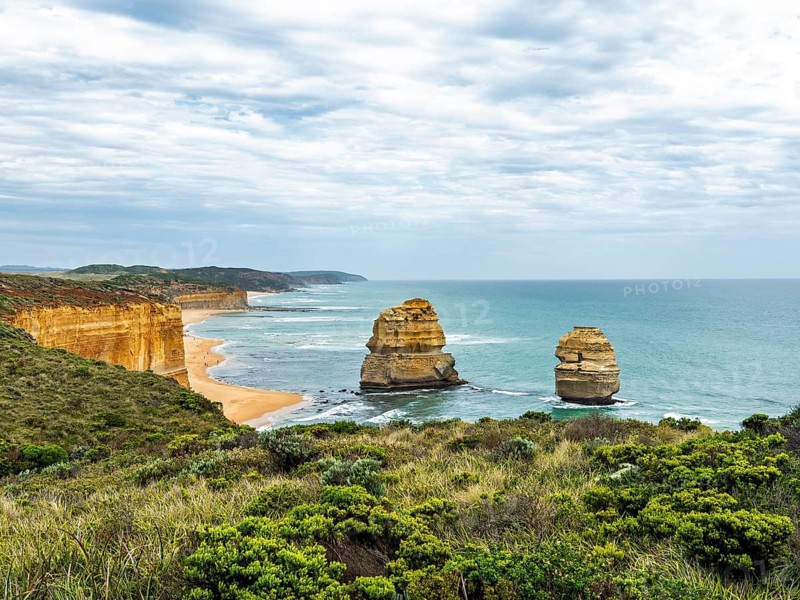 Dramatic coastal rock formations rise from the turquoise ocean, bordered by lush plant life and sandy cliffs under a partly cloudy sky in the Twelve Apostles, Australia