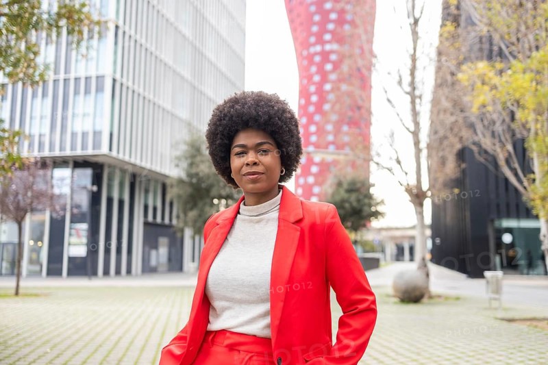 A confident woman in a vibrant red suit stands in an urban setting, exuding style and professionalism. Modern architecture and natural elements provide a dynamic backdrop