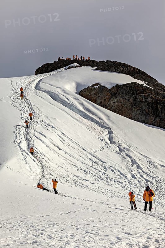 People in orange jackets climb a snow-covered mountain, tourists hike across the ice and rocky landscape of Antarctica
