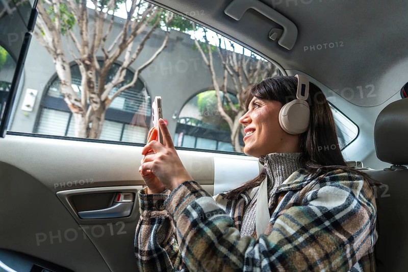 Woman wearing headphones and plaid jacket riding in the back seat of a car, engaging with her smartphone and enjoying the urban scenery during a city trip