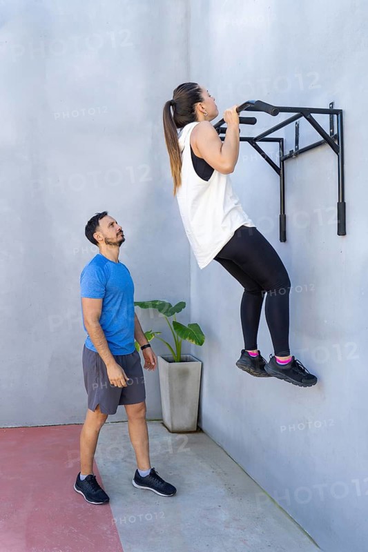 Determined woman performing pull-ups on a wall-mounted bar while personal trainer observes and supports, creating a motivating training environment