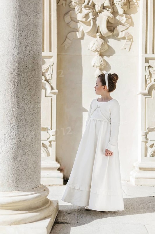 A young girl stands gracefully in a First Communion dress, adorned with a headband, beside ornate architectural details. The setting exudes elegance and tradition
