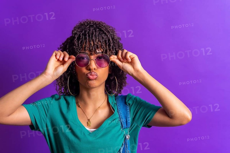 Fashionable young woman with curly hair adjusting her purple sunglasses and pouting on a vibrant purple background
