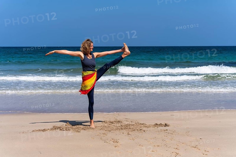 A woman doing yoga on a sandy beach by the sea. Vrksasana, tree, variant. Treen Cove, Cornwall, England, Great Britain