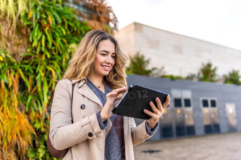 Young woman dressed in a trench coat smiling and interacting with a digital tablet, conducting business or connecting using wireless technology in an urban setting with a green wall