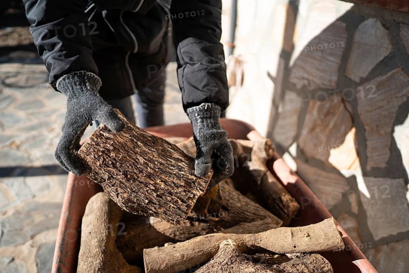 A cropped unrecognizable person places logs into a wheelbarrow, preparing them for a wood burning stove. He's wearing gloves and involved in outdoor activity, unrecognizable from this perspective