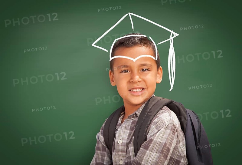 Young Hispanic Student Boy Wearing Backpack Front Of Blackboard with Graduation Cap Drawn In Chalk Over Head