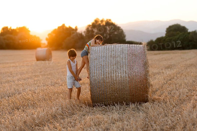 Two siblings, a boy and a girl, play near a large haystack in a field bathed in golden sunset light, conveying a sense of rural adventure and childhood joy