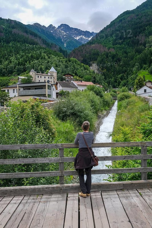 Tschengls, Vinchgau, South Tyrol, Italy, Mountain landscape behind the village of Tschengls, Hiker stands on a wooden bridge over a mountain stream and looks towards Tschenglsburg and mountain landscape