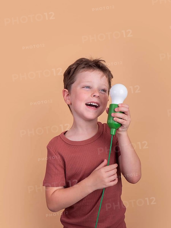 A joyful child in a red shirt holds a green handled light bulb against a peach background, exuding curiosity and creativity. Perfect for themes of innovation and imagination