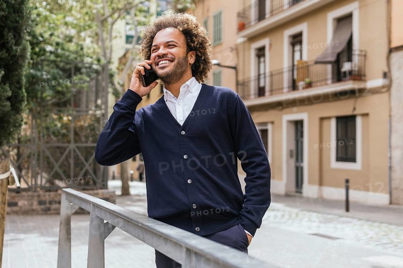 A smiling man takes a break from work at a coworking space in Barcelona, using his smartphone outside. The scene captures a moment of relaxation amidst urban architecture