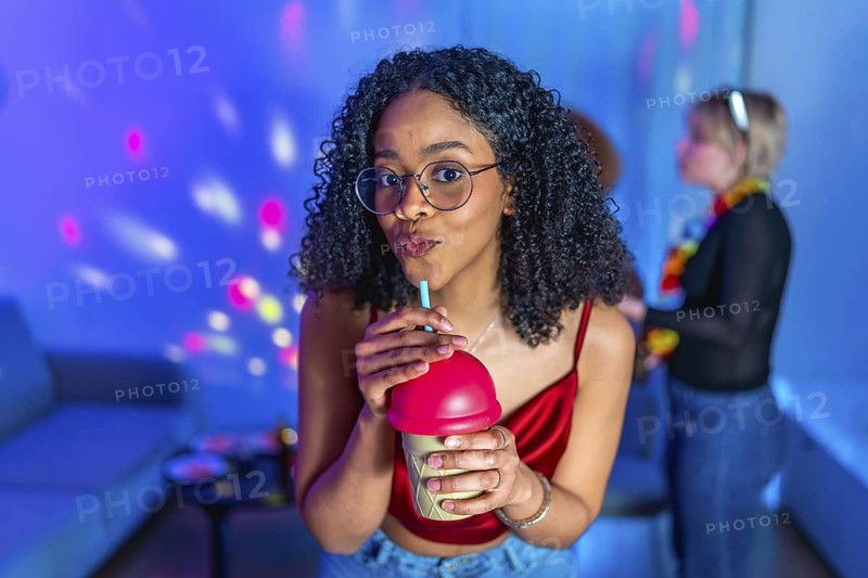 Young woman sipping a beverage through straw, enjoying a vibrant karaoke night with friends, illuminated by colorful disco lights
