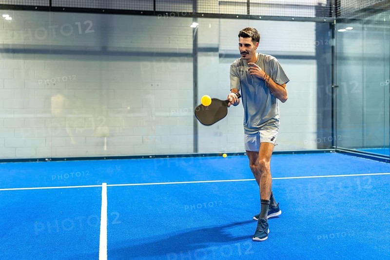 Man playing padel on a vibrant blue court, holding a paddle racket and hitting a bright yellow pickleball during an intense match, showcasing action and sports performance