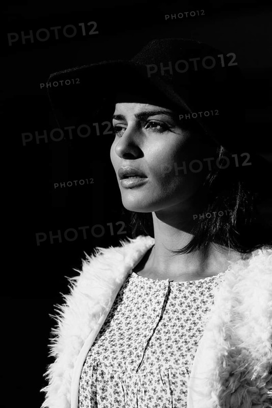 A black and white portrait of a woman in a wide-brimmed hat. The dramatic lighting and shadows emphasize her serene expression and elegant attire