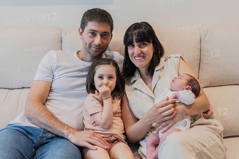 A young family sitting together, showcasing love and happiness with their newborn baby in the mother's arms, while the big sister sits playfully beside the father