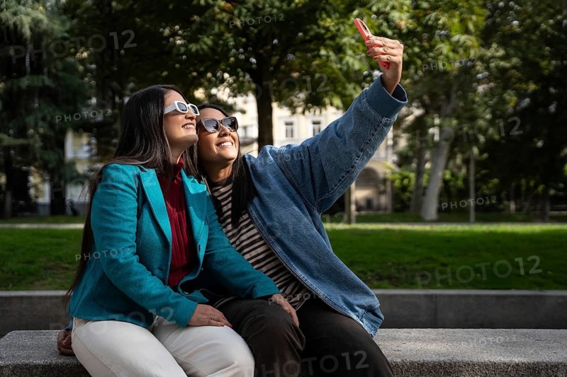 A joyful Latin lesbian couple in sunglasses takes a selfie in a sunny park. They are sitting on a bench, embracing and smiling, surrounded by lush greenery and trees