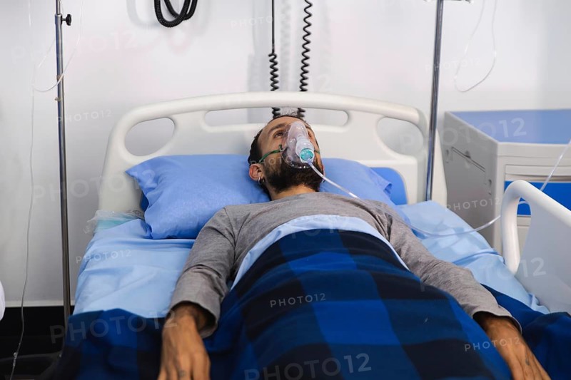 A patient lies in a hospital bed using an oxygen mask for respiratory support. Various medical devices and IV lines are visible, illustrating a clinical healthcare environment
