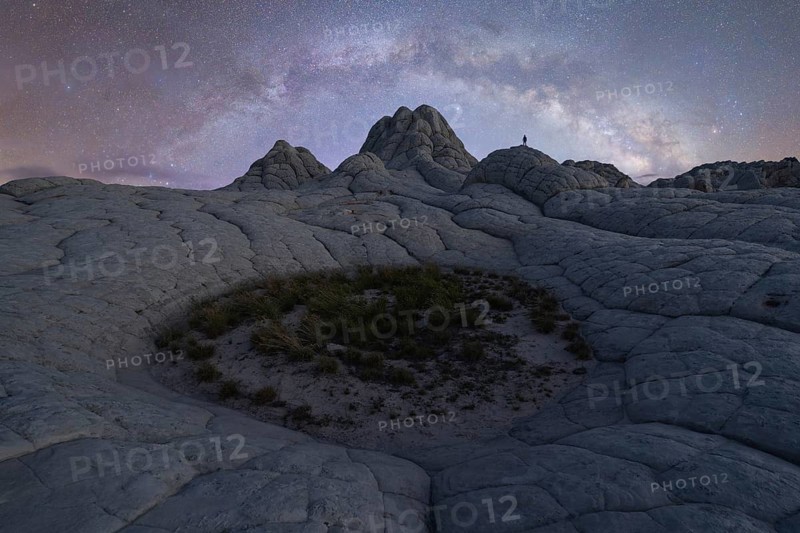 A lone man stands beneath the sprawling Milky Way in the otherworldly terrain of White Pocket, Arizona. The night sky illuminates the desert landscape below