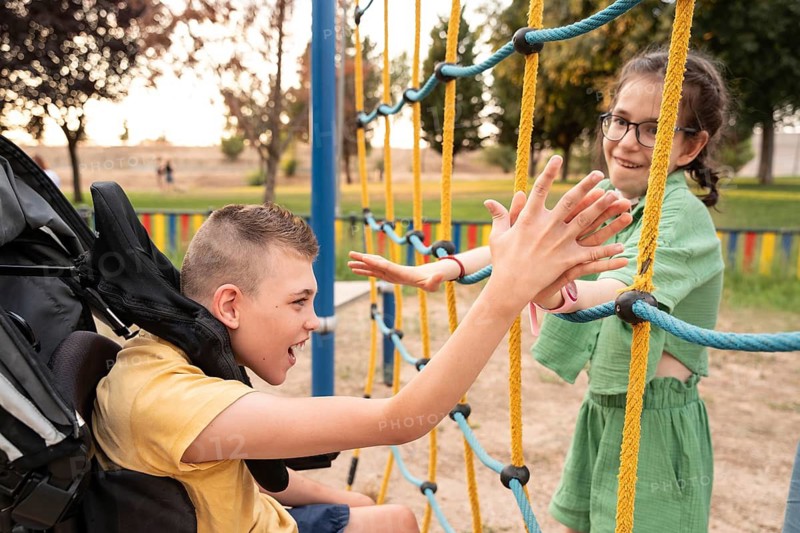 A boy with cerebral palsy and a girl with a tracheostomy enjoy a playful moment on a rope climbing structure in a park, highlighting friendship and inclusion