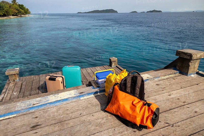 A collection of travel bags sits on a wooden dock, with crystal clear tropical waters and distant islands in the background, evoking a sense of adventure and relaxation in Indonesia
