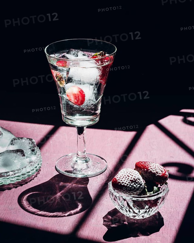 A refreshing gin tonic with ice cubes and strawberries sits elegantly on a pink table. Sunlight and shadows create a vibrant and inviting summer cocktail scene