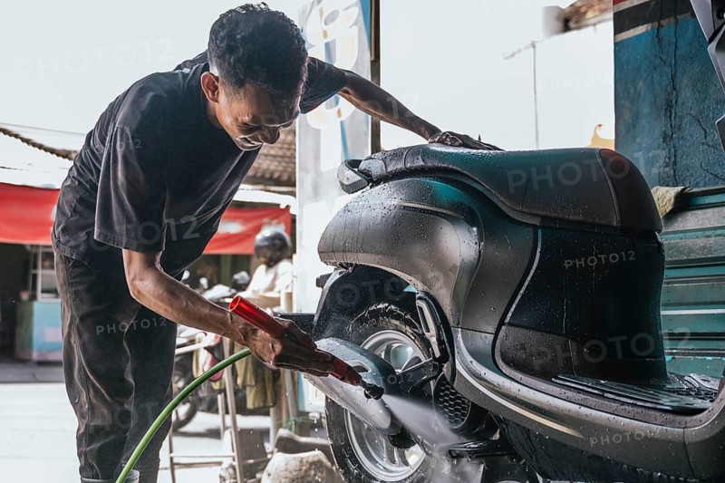A professional cleaner thoroughly washes a motorcycle using a water hose, focusing on the wheel and body. The scene captures the dedication to detail and care in vehicle maintenance