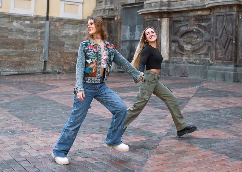 Two young woman friends energetically walk hand in hand across a brick paved area, exuding joy and friendship. The city ambiance adds to their carefree moment
